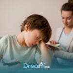 Autistic boy looking frustrated while drawing, resting head on hand as adult in background observes & provides gentle support
