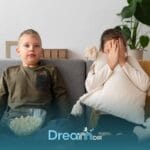 Two autistic kids sitting on couch watching TV, one calmly eating popcorn while the other hides face with pillow