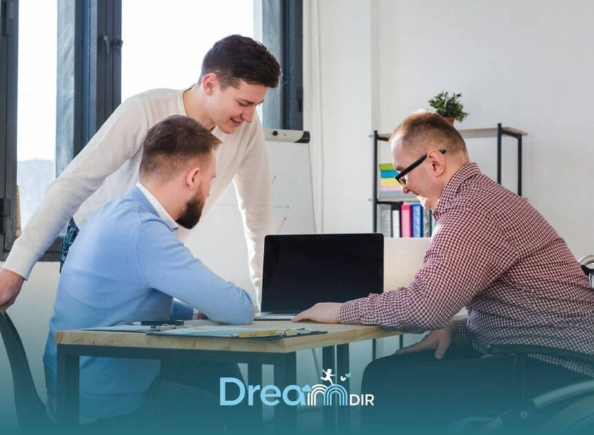 Three autistic adults collaborating at desk with laptop, reviewing work together in a supportive & inclusive workplace.