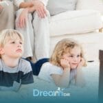 Two young autistic children lie on a soft carpet watching television together while parents relax in the background at home