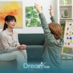 Autistic boy raises hands during a sensory integration therapy activity while therapist sits nearby during therapy
