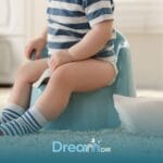 Autistic boy wearing striped socks sits on a potty training chair beside a roll of toilet paper in a bright home setting