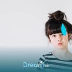 Autistic young girl with a thoughtful expression stands against a plain background with a blue masking symbol beside her face